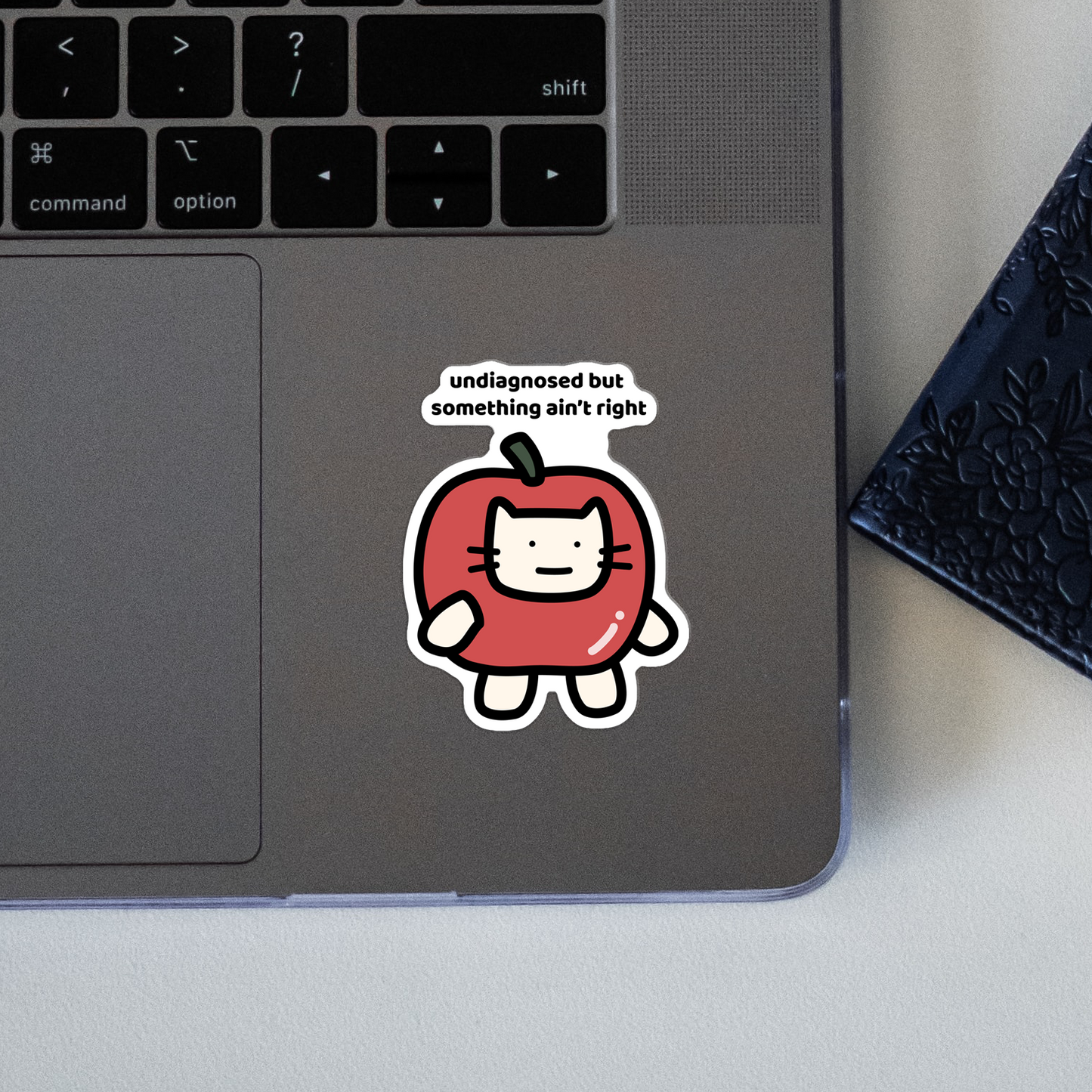 closeup of waterproof apple cat vinyl sticker with the text "Undiagnosed but something ain't right"