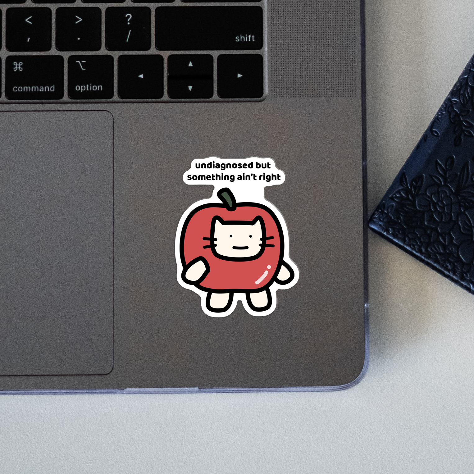 closeup of waterproof apple cat vinyl sticker with the text "Undiagnosed but something ain't right"