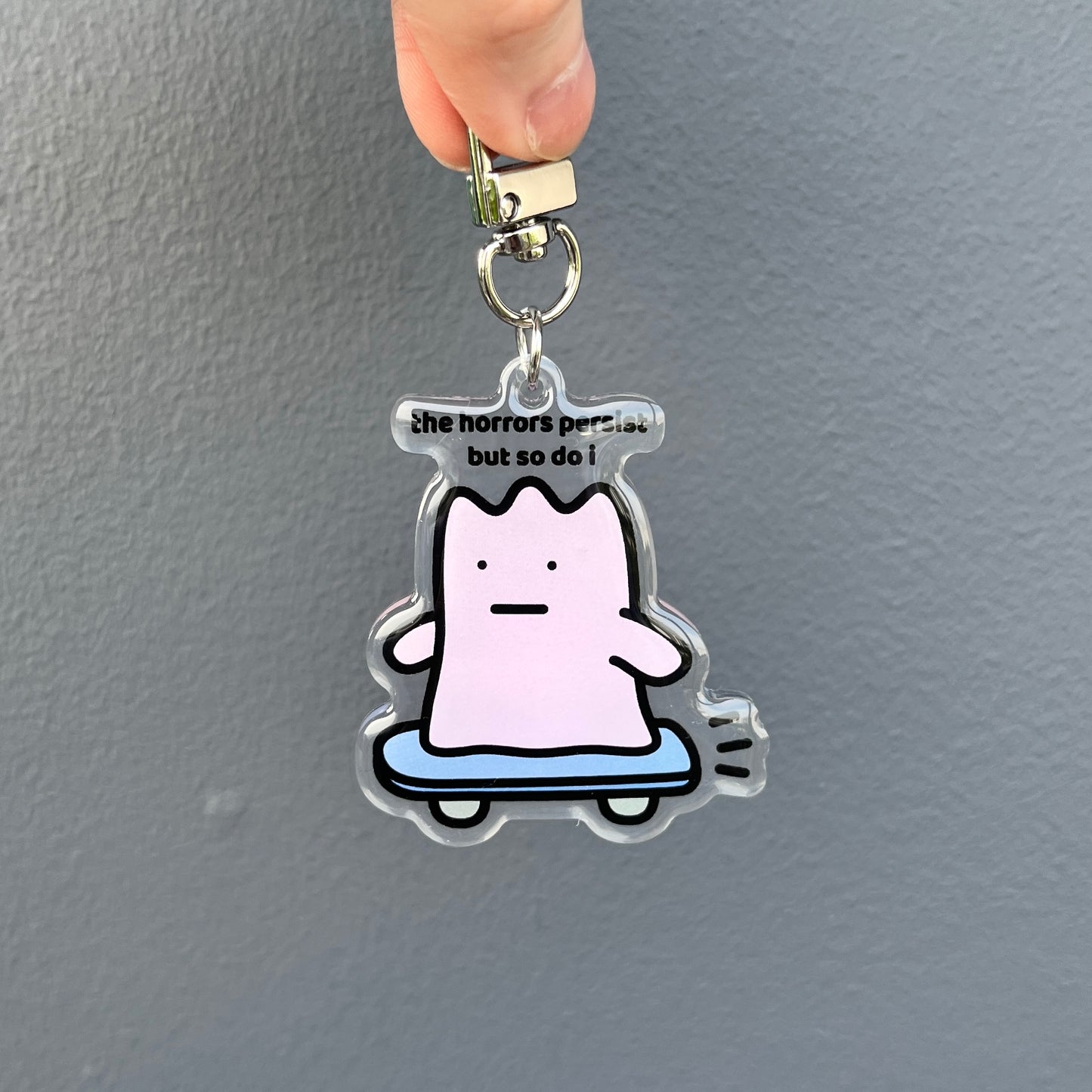waterproof skateboard acrylic keychain with the text “The horrors persist but so do I" on a plain background